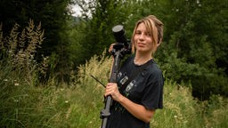Die ukrainische Filmemacherin Anja Korman-Lozinsk posiert mit einer Kamera vor einer grünen Wald- und Wiesenlandschaft.