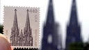 Eine Briefmarke mit dem Kölner Dom wird am 10.8.2001 vor dem Original hochgehalten.