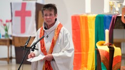Gemeindereferentin Marianne Arndt predigt bei einer Messe, eine Regenbogenflagge hängt im Hintergrund.