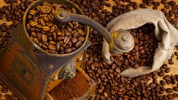 Nahaufnahme einer mit Kaffebohnen gefüllten Kaffeemühle, daneben ein Sack voller Kaffebohnen.