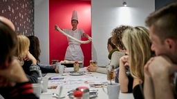 Teilnehmer:innen der Kulinarischen Weltreise des Fremdenverkehrsmtes "Kulturklüngel" sitzen an einem Tisch und schauen einem Koch zu.