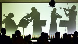 Die Silhouetten von live spielenden Orchestermusikern sind auf einer Leinwand zu sehen, davor die Silhouetten des Publikums.