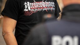 Ein Teilnehmer einer Demonstration trägt ein T-Shirt mit der Aufschrift "Ureinwohner", während er von einem Polizisten kontrolliert wird.