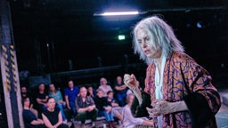 Szenenfoto von "Aufzeichnungen aus dem Kellerloch" bei der Ruhrtriennale: Eine Frau (Nina Hoss) mit längeren grauen Haaren gestikuliert vor Menschen im Publikum.