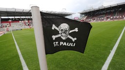 Eine Totenkopfflagge mit dem Schriftzug "St. Pauli" flattert vor einem Spiel im Millerntor-Stadion an einer Eckfahne im Wind.