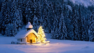 Schneebedeckte Kapelle mit beleuchtetem Tannenbaum daneben
