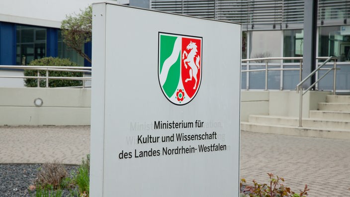 Außenansicht des Geländes sowie ein Hinweisschild mit dem Schriftzug "Ministerium für Kultur und Wissenschaft des Landes Nordrhein-Westfalen" und Landeswappen.