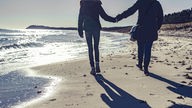 Silhouette von zwei Frauen, die Hand in Hand am Strand spazieren gehen.