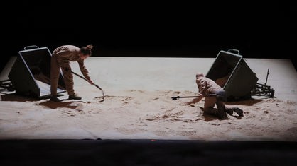 Zwei Personen arbeiten auf einem sandigen Boden mit Werkzeugen.