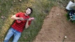 Ein Junge (Mike) liegt neben einem Grabstein und guckt in den Himmel, er spielt dabei auf einer Ukulele.