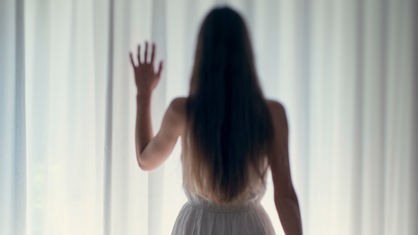 Das Bild ist verschwommen, eine Frau in einem Kleid steht vor einem mit Vorhang zugezogenem Fenster.