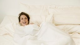 Ein Paar im Bett, die Frau lacht während der Mann unter der Decke verschwindet.