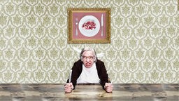 Ein Mann mit grauem Haar und Brille am Tisch, hält Messer und Gabel in den Händen, dahinter ein Foto mit Fleisch an der Wand.