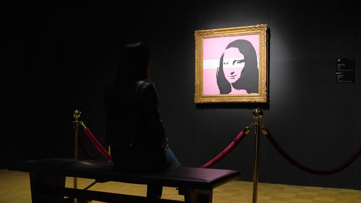 Das berühmte Mona Lisa-Bild mit weiß-rosa Streifen von Banksy bearbeitet, erleuchtet an einer sonst dunklen Wand.