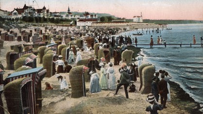 Die colorierte Postkarte aus der Zeit um 1910 zeigt das Strand- und Badeleben am Strand des Ostseebades Binz auf der Insel Rügen.