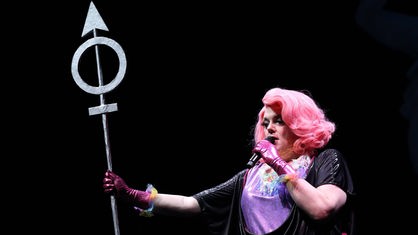 Schauspieler:in Kaey Kiel steht für das Theaterstück "Planet Egalia" auf der Bühne. Sie trägt eine pinke Perücke und Glitzerkleidung, in der Hand hält sie ein Schild in Form des Marssymbols. 