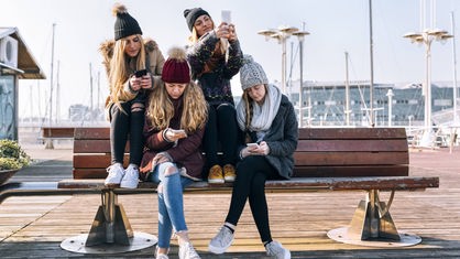 Vier junge Mädchen sitzen auf einer Bank und spielen mit ihrem Handys.