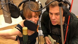 Mann und Frau mit Kopfhörern im Studio vor Mikros am singen, er spielt zusätzlich Gitarre.