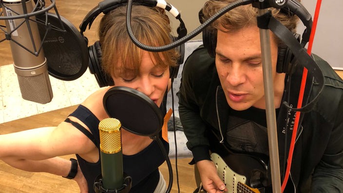 Mann und Frau mit Kopfhörern im Studio vor Mikros am singen, er spielt zusätzlich Gitarre.