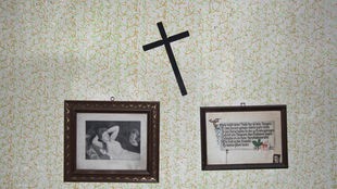 Ein schiefes Kreuz und zwei alte Bilder hängen an einer Wand.