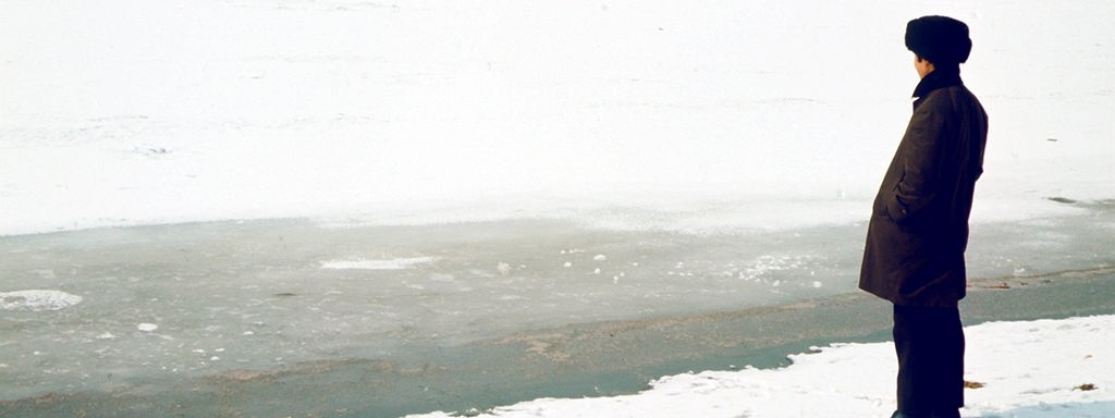 Ein Mann mit Pelzmütze und Winterkleidung steht am Ufer des zugefrorenen Amur nahe Chabarowsk und schaut in Richtung einer Gruppe Eisfischer.