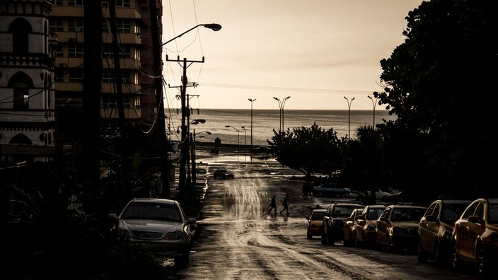 Eine Straße in Havanna, Kuba führt zum Meer, an der Seite parken Autos.