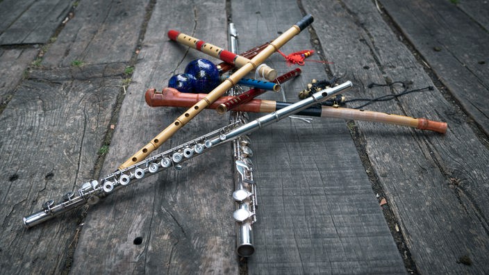 Verschiedene Flöten liegen quer übereinander gestapelt auf alten Holzbohlen.