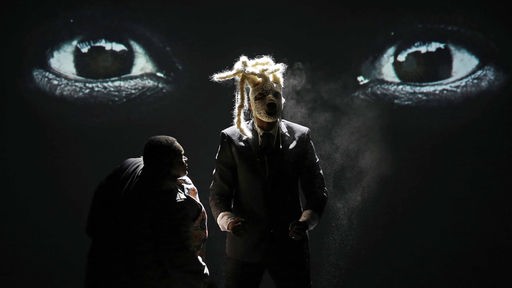 Szenenfoto der Produktion "Samson": Eine Mann blickt auf eine Person mit einer weißen Maske, im Hintergrund ein überdimensionales Augenpaar.