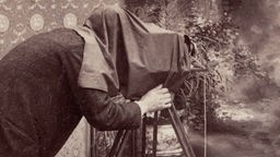 Eine Sepia-Fotografie eines Fotografen unter einem schwarzen Tuch mit einer Plattenkamera.