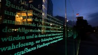 Textauszug des Grundgesetzes auf einer Glaswand, im Hintergrund der Deutsche Reichstag mit einer Deutschland-Fahne in der Dämmerung.