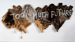 Ausstellungsansicht, Werk von Rebekka Benzenberg: Nebeneinander gehängte Pelzmäntel und mit der Aufschrift "Too Much Future".