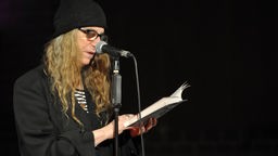Patti Smith mit dunklem Sacke und Wollmütze liest hinter einem Mikrofon stehend aus ein Buch (Symbolbild).