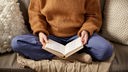Eine Frau sitzt mit einem warmen Wollpullover auf dem Sofa und liest ein Buch.