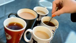 Ein Hand rührt in einer von mehreren nebeneinanderstehenden, mit Kaffee oder Tee gefüllte Tasse.