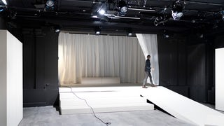 Eine Person öffnet den weißen Bühnenvorhang in einem schwarz gestrichenen Theatersaal