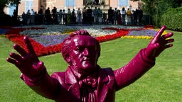 Lila gefärbte Plastik-Figur, die den Komponisten Richard Wagner darstellt, im Hintgrund Besucher auf dem Hügel der Bayreuther Festspiele.