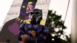Ein dunkelhäutiges Kind, im Kostüm eines Superhelden aus dem Film Black Panther, steht vor einem Wandgemälde eines schwarzen Superhelden