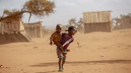 Ein kleines Mädchen trägt unter großer Anstrengung ein Kleinstkind durch eine dürr-staubige Steppenlandschaft, im Hintrgrund sind Hütten zu sehen.