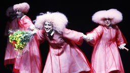 Szenefoto von "Bühnenbeschimpfung": Mehere weiß geschminkte Personen in rosa-farbenen Kleidern und Perücken halten sich an den Händen.