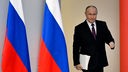 Der russische Präsident Wladimir Putin mit Papieren im Arm auf dem Weg zu seiner Rede zur Lage der Nation, im Hintergrund Russland-Flaggen.