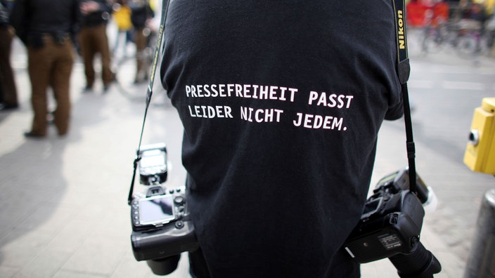 Ein:e Fotograf:in trägt eine Jacke mit dem Aufdruck: "PRESSEFREIHEIT PASST LEIDER NICHT JEDEM."