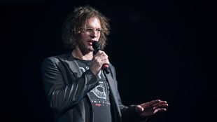 Tim Isfort spricht mit einem Mikrofon in der Hand auf einer dunklen Bühne.