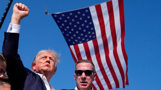 Donald Trump leicht am Ohr blutend, ballt die Faust, neben ihm Agenten des US-Geheimdienstes Secret Service umringt, darüber die Flagge der USA.