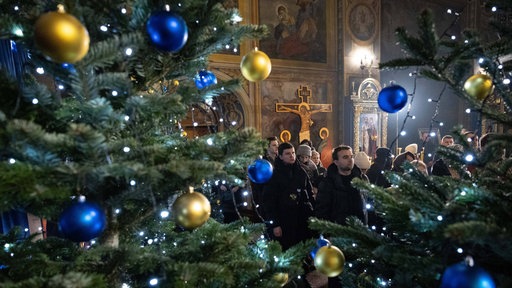 Symbolbild von 2023: Heiligabend-Gottesdienst der ukrainisch-orthodoxen Kirche in Kiew: Weihnachtsbäume mit Lichterketten, gelben und blauen Kugeln, im Hintergrund Menschen.