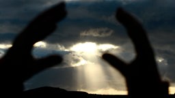 Hände versuchen eine von Wolken teilweise verdunkelte Sonnen einzurahmen.