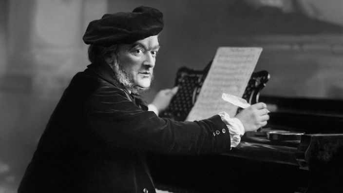 Ein Schwarz-Weiß Portrait des Komponisten Richard Wagner, am Klavier sitzend. Er sitzt mit einer Schreibfeder in der Hand vor seinen Noten.
