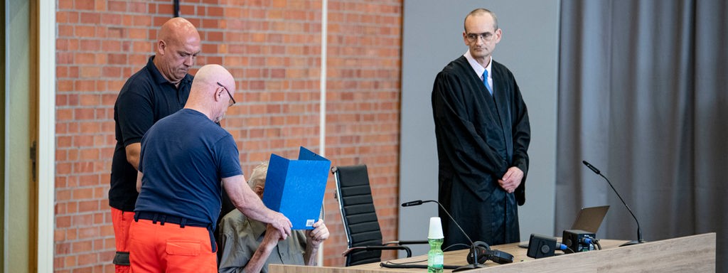 Der Angeklagte verdeckt sein Gesicht mit einer blauen Mappe, links von ihm assistieren zwei Sanitäter, rechts steht sein Anwalt.