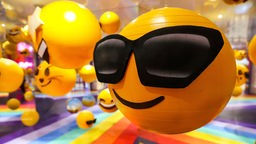 Ein gelber, runder Emoji, der eine Sonnenbrille trägt. 