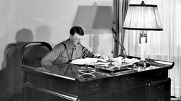 Eine Schwarz-Weiß Aufnahme von Hitler an seinem Schreibtisch, wie er in einem Buch liest.