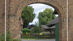 Kulturhof Velbrück: Blick durch einen gemauerten Torbogen auf ältere Backsteingebäude neben großen Bäumen.